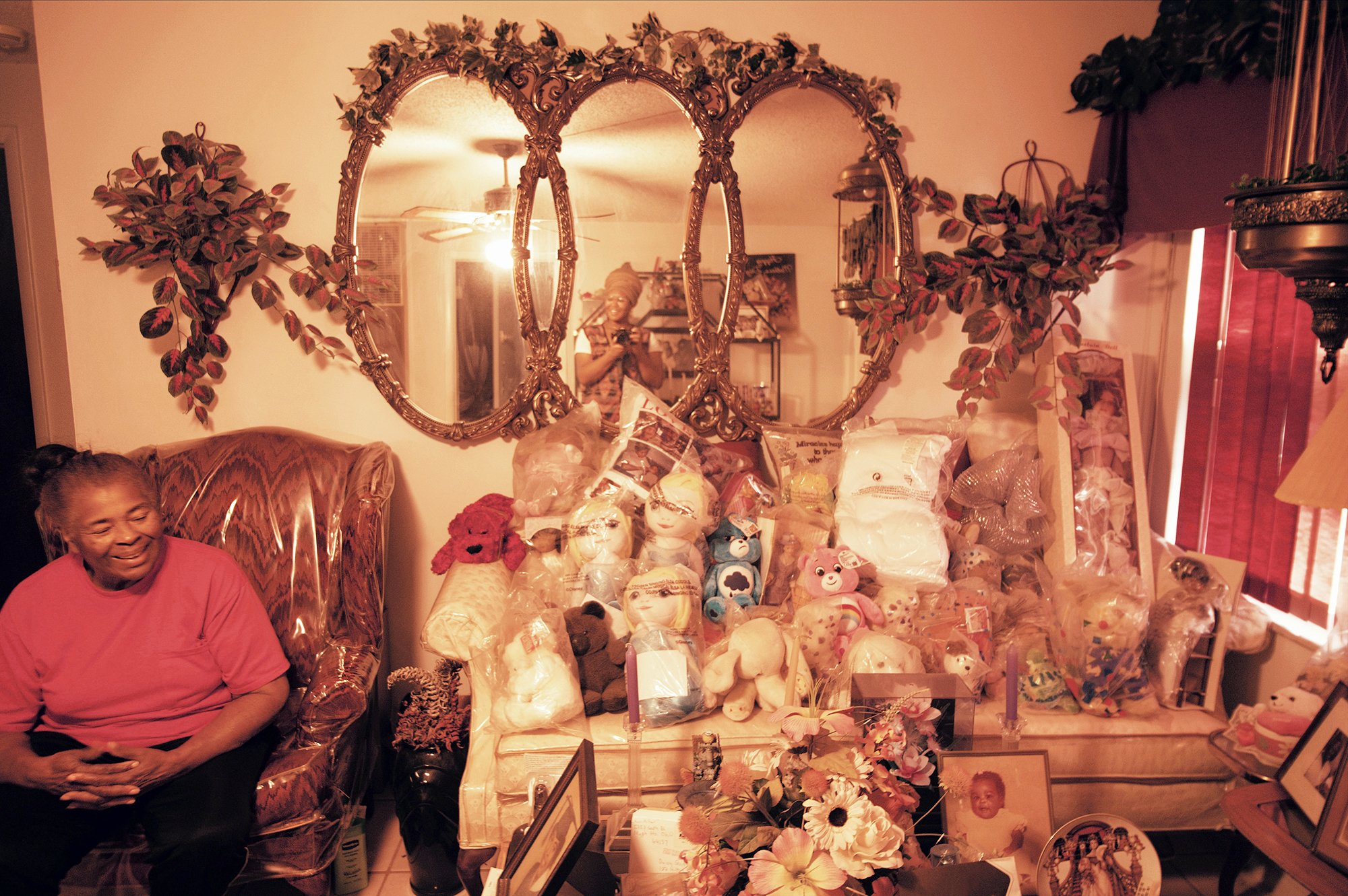 The lady sitting in a chair next to a sofa filled with soft toys, a large mirror piece on the wall.