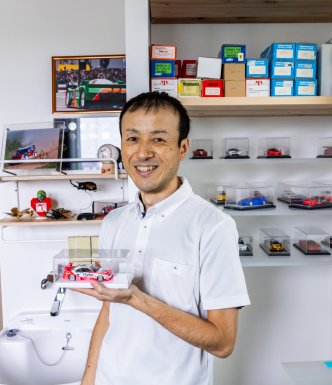 Man smiling, holding a model car, with other model cars arranged neatly on a shelf behind him