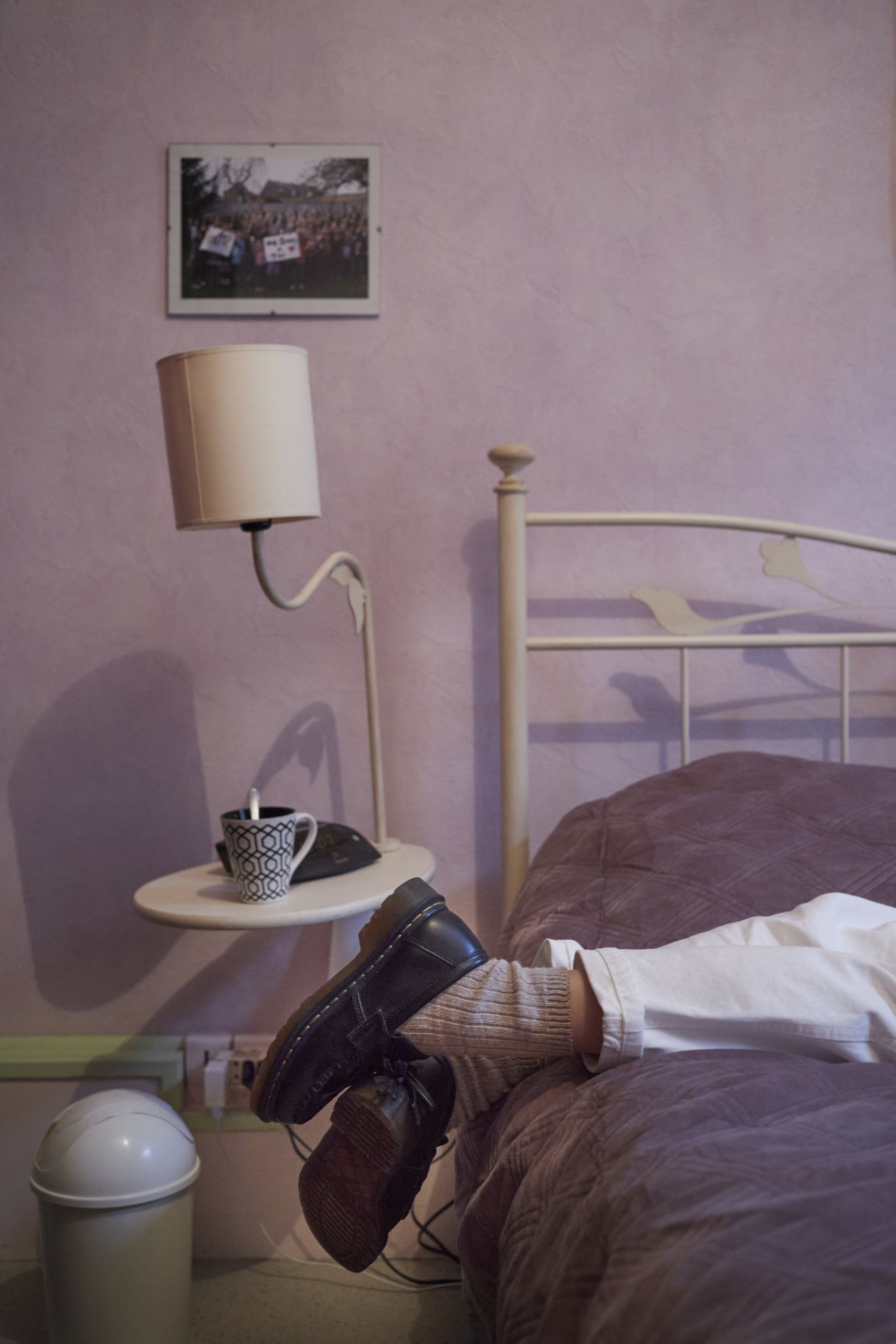 A persons legs on a bed in a purple bedroom, the person is wearing white pants and black shoes.