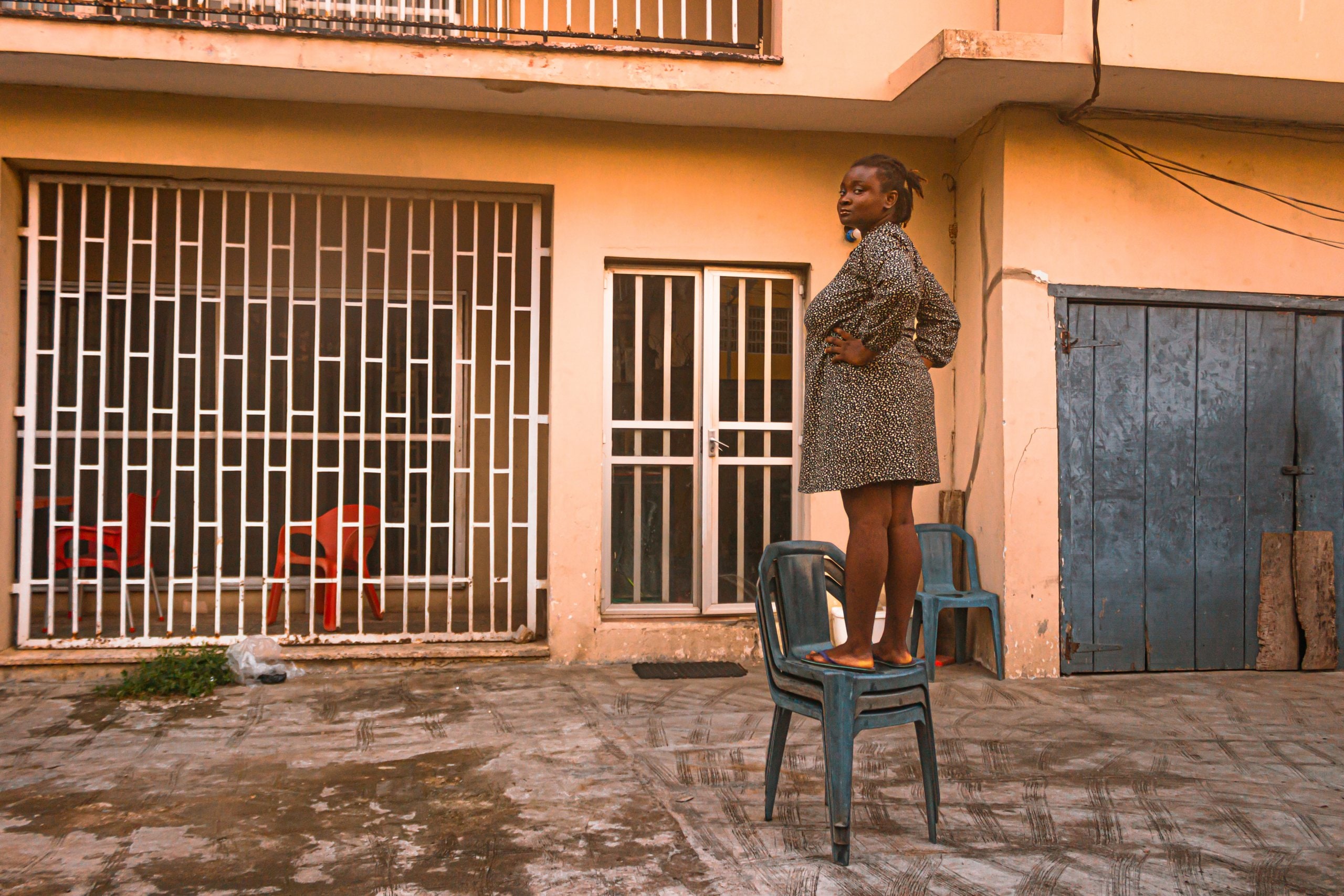 Ife standing on three stacked plastic chairs outside her house.