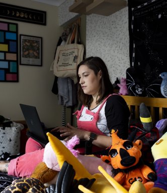 Lady sitting on her bed, using her laptop, surrounded by plush toys