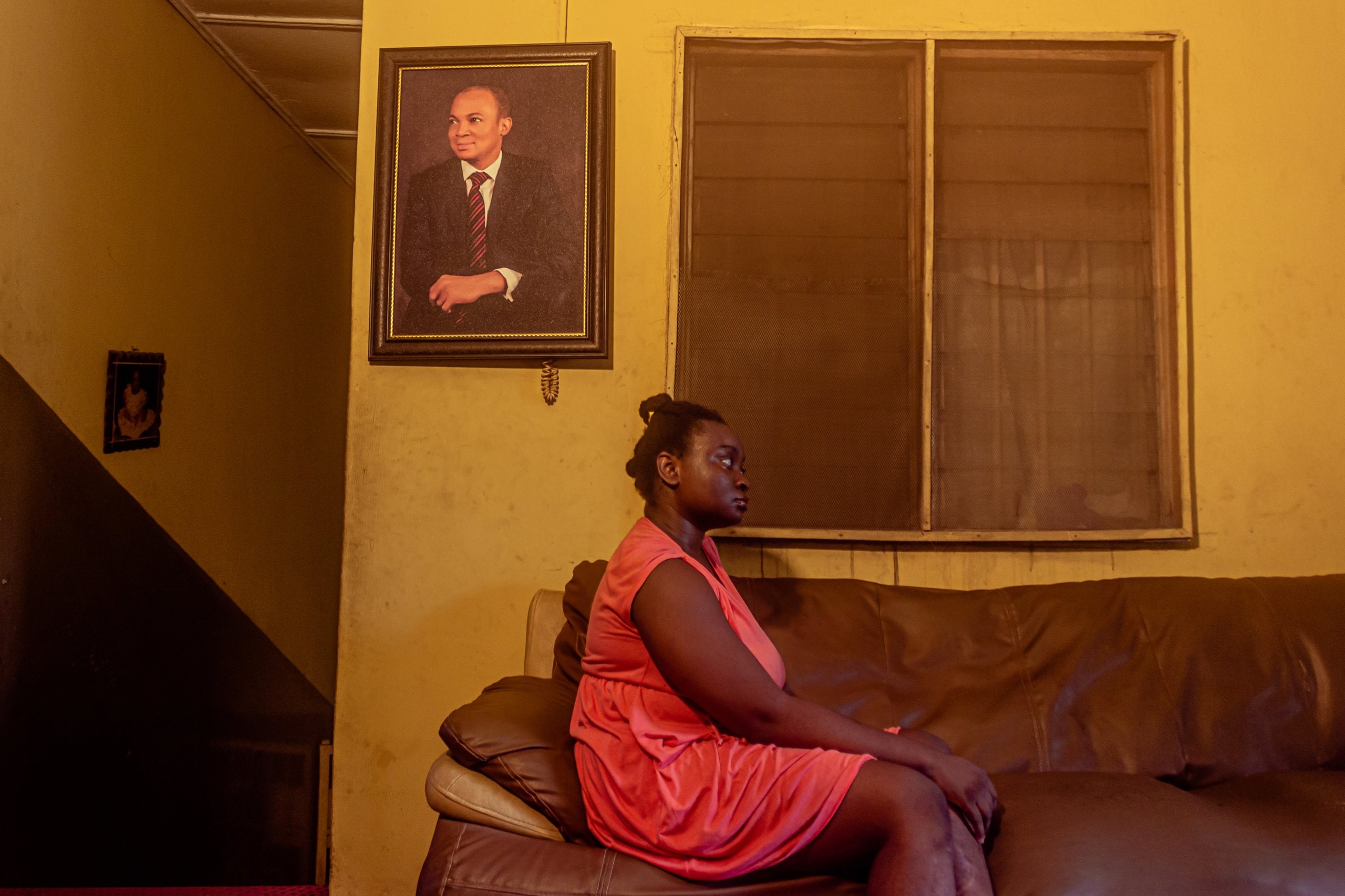 Ife sitting on a sofa in front of a window, a photo of a man in a suit on the wall behind her.