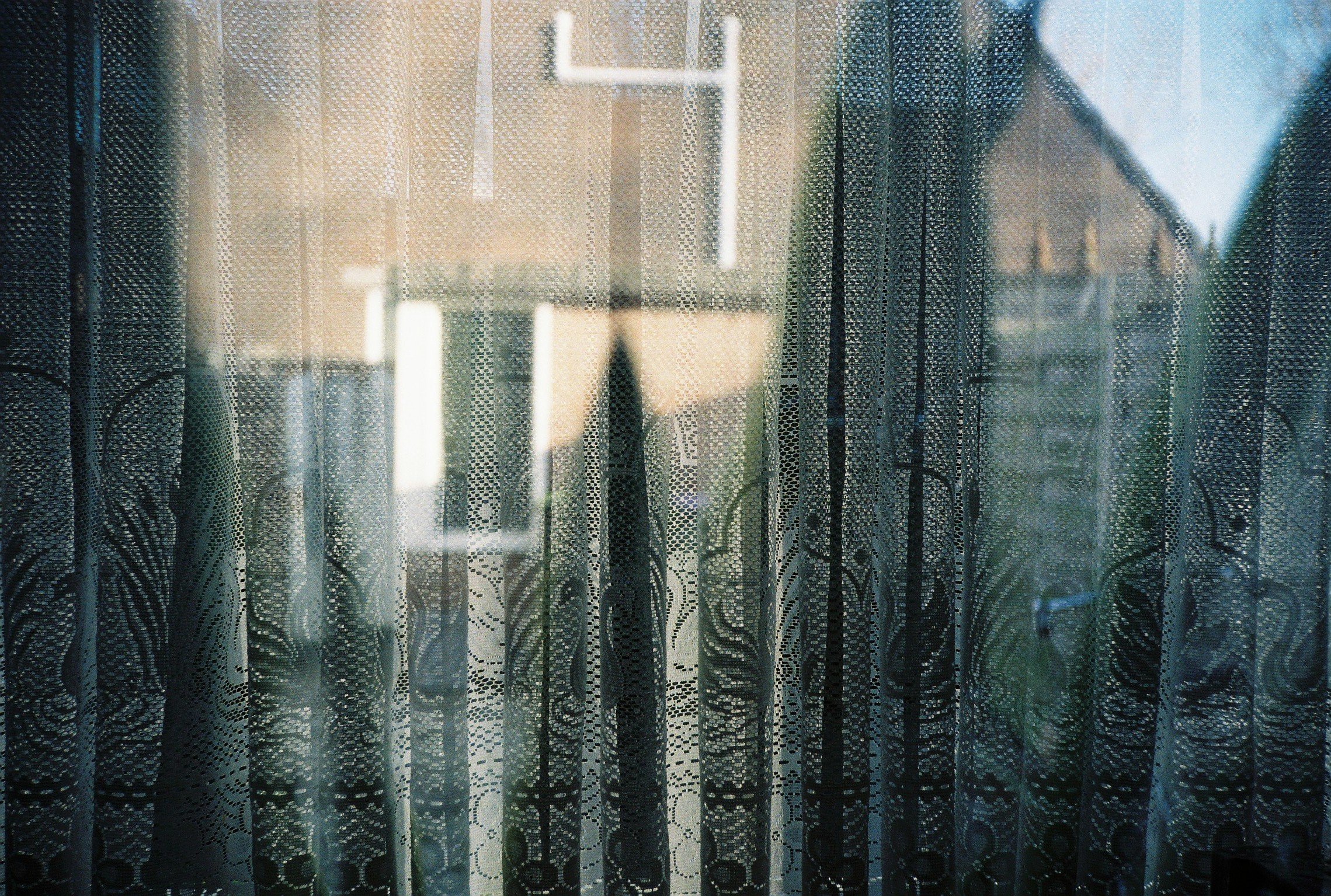 Close-up of a window with lace curtains, a house outside the window.